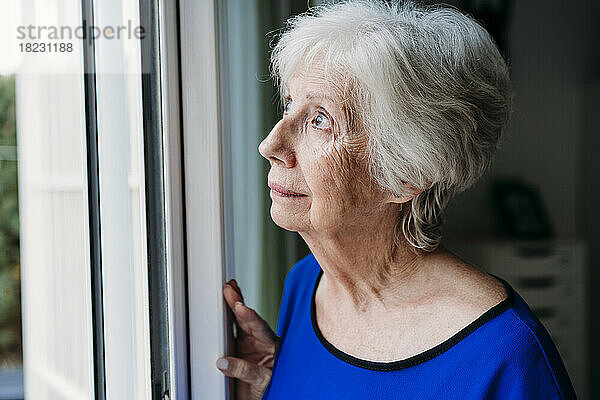 Nachdenkliche ältere Frau schaut zu Hause durch das Fenster