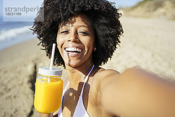 Glückliche Frau  die im Sommer ein Selfie mit Orangensaft am Strand macht