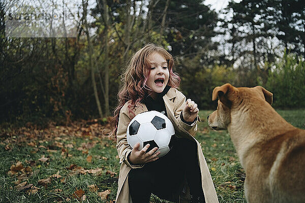 Nettes Mädchen hält Fußball und spielt mit Hund im Park