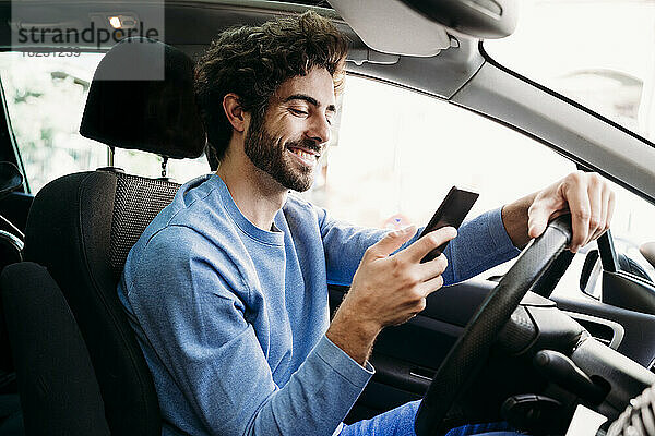 Lächelnder Mann sitzt auf dem Fahrersitz im Auto und benutzt ein Smartphone
