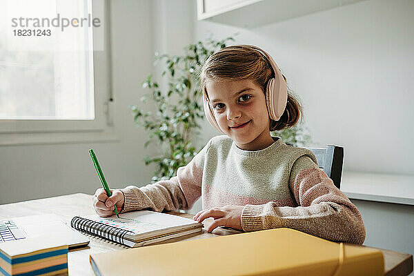 Lächelndes Mädchen mit kabellosen Kopfhörern  das zu Hause am Tisch lernt