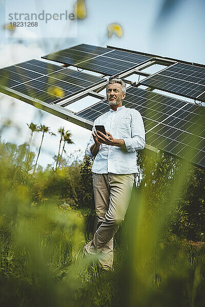 Reifer Mann stützt sich mit Smartphone im Garten auf Sonnenkollektoren