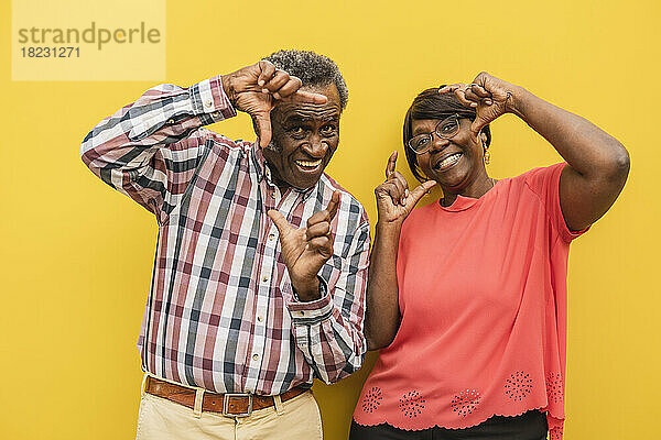 Glückliches Seniorenpaar macht Fingerrahmen vor gelbem Hintergrund
