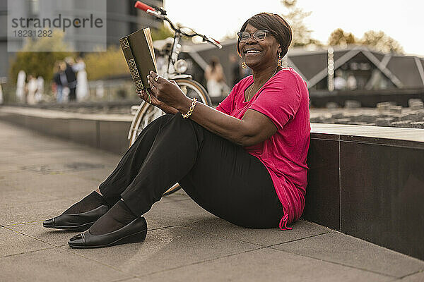 Lächelnde Frau  die auf dem Fußweg ein Buch liest