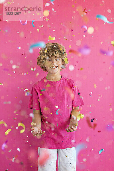 Fröhlicher Junge spielt mit Konfetti vor rosa Hintergrund