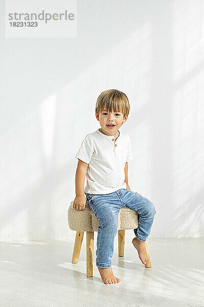 Cute boy sitting on stool in front of wall at home