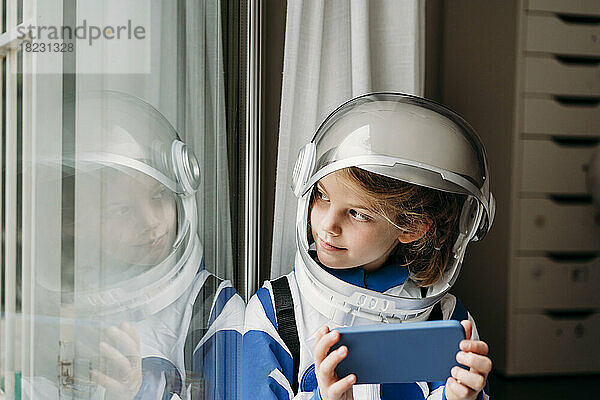 Lächelndes Mädchen im Weltraumkostüm mit Mobiltelefon und Blick auf die Spiegelung am Fenster