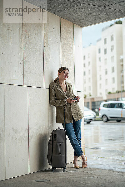 Lächelnde Geschäftsfrau lehnt mit Gepäck an der Wand