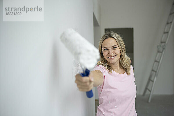 Lächelnde Frau hält Farbeimer in der Nähe der weißen Wand in der Wohnung