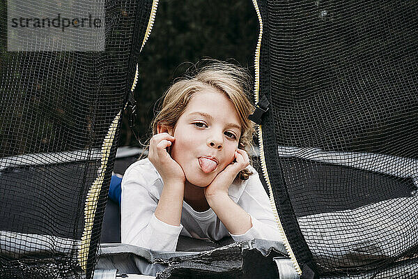 Mädchen streckt Zunge heraus und liegt auf Trampolin im Garten