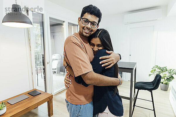 Lächelnder junger Mann umarmt glückliche Frau im heimischen Wohnzimmer