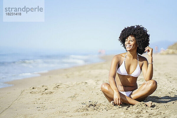 Glückliche junge Frau sitzt im Sommer auf Sand am Strand