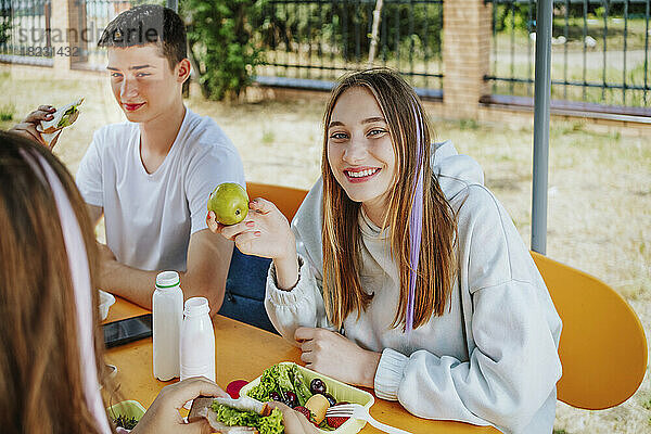 Glückliches Mädchen  das frisches Obst isst und am Tisch auf dem Schulhof sitzt