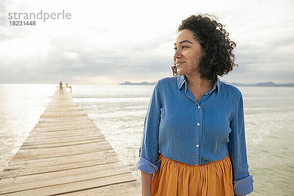 Lächelnde Frau mit lockigem Haar steht auf einem Steg am Meer