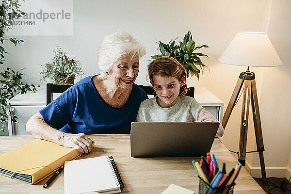 Lächelnde ältere Frau hilft ihrer Enkelin beim Lernen per Laptop auf dem heimischen Tisch