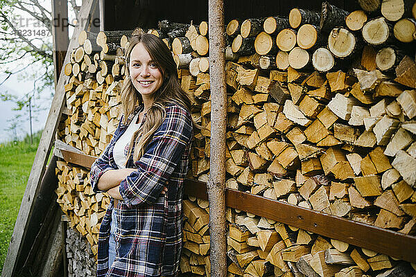 Lächelnde junge Frau mit verschränkten Armen steht vor Baumstämmen