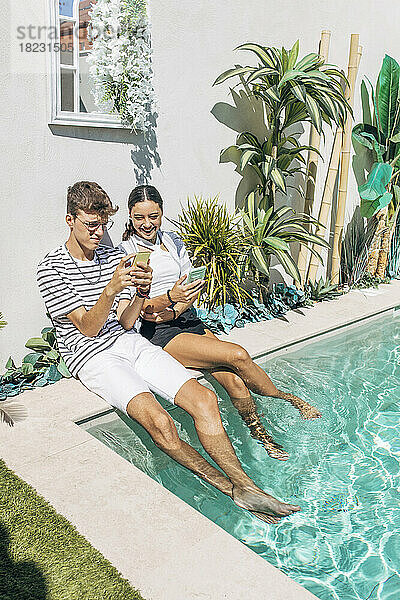 Lächelndes Mädchen und Junge  die an einem sonnigen Tag ihr Smartphone am Schwimmbad benutzen