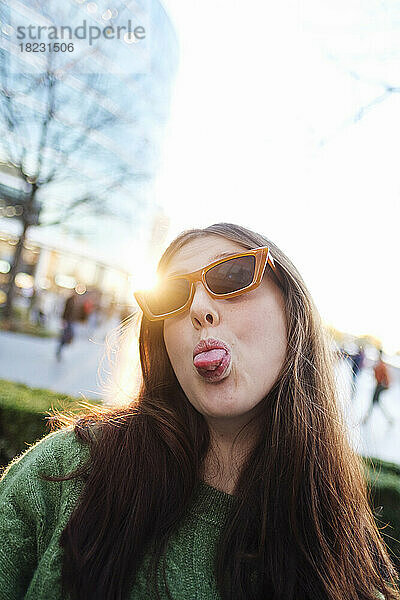Junge Frau mit Sonnenbrille streckt vor strahlend klarem Himmel die Zunge heraus