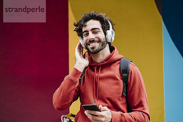Glücklicher junger Mann mit kabellosen Kopfhörern vor bunter Wand