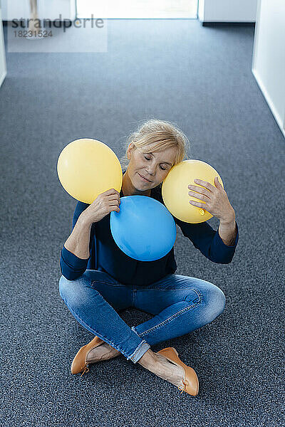 Geschäftsfrau mit geschlossenen Augen und Luftballons  die im Büro auf dem Boden sitzt