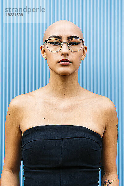 Selbstbewusste junge Frau mit rasiertem Kopf und Brille vor blauer Wand