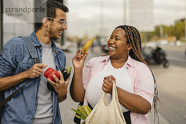 Fröhlicher junger Mann mit Freundin hält eine Tüte Gemüse am Fußweg