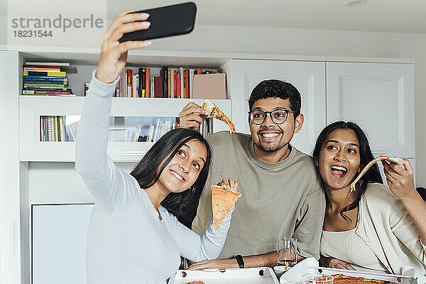 Lächelnde Frau macht Selfie per Smartphone  während Frau und Mann zu Hause ein Pizzastück zeigen