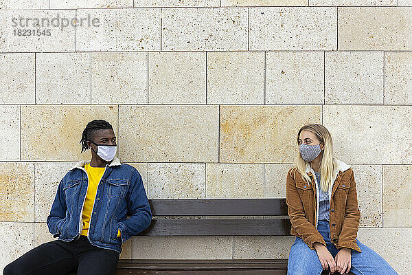 Junges Paar mit Schutzmasken sitzt auf Bank