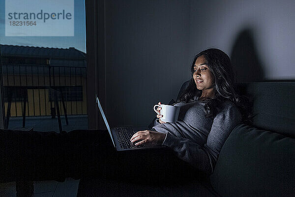Lächelnde junge Frau mit Kaffeetasse und Laptop zu Hause