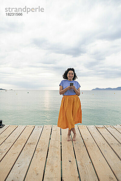Lächelnde Frau steht mit Smartphone am Steg vor dem Meer