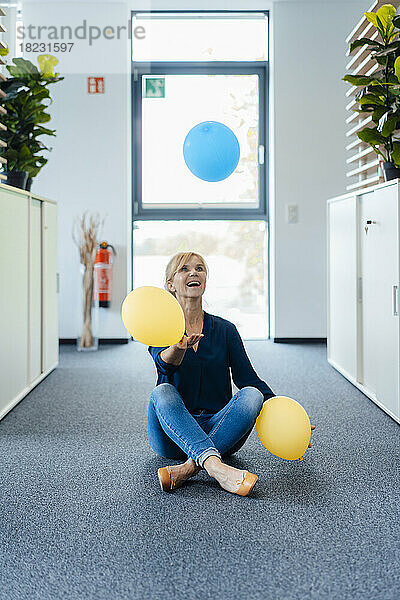 Happy businesswoman playing with balloons sitting on ground at workplace