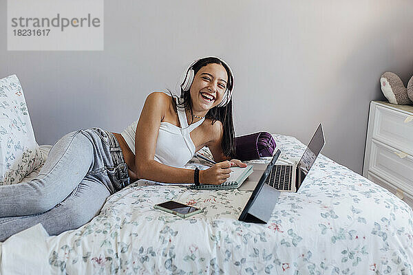 Fröhliches Mädchen trägt Kopfhörer und macht Notizen mit Gadgets im Schlafzimmer