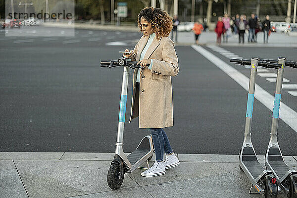 Junge Frau mietet Elektroroller über Smartphone auf Fußweg