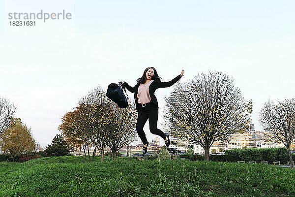 Glückliche junge Frau mit Tasche springt im Park