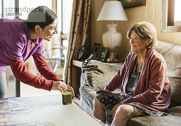 Glückliche Pflegekraft rührt Kaffee um und spricht mit einer älteren Frau  die zu Hause auf dem Sofa sitzt