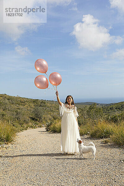 Fröhliches Mädchen in weißem Kleid hält Luftballons neben Hund auf dem Weg