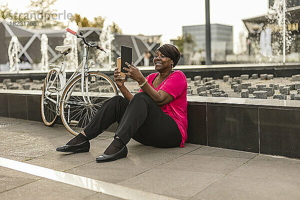 Lächelnde Frau sitzt mit dem Fahrrad in der Nähe der Wand und benutzt einen Tablet-PC