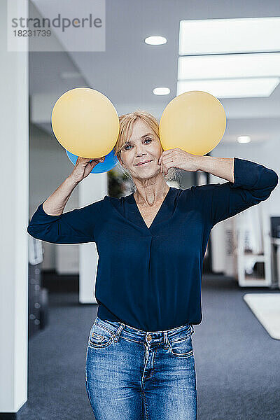 Lächelnde reife Geschäftsfrau hält Luftballons am Arbeitsplatz