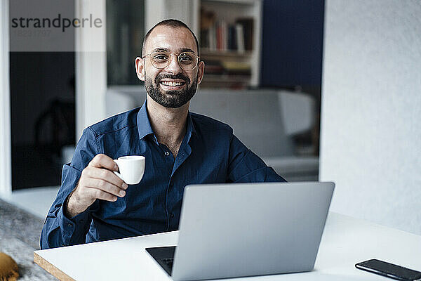 Happy businessman holding coffee cup sitting with laptop at desk in office