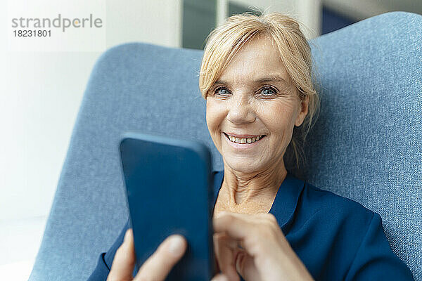 Glückliche reife Geschäftsfrau mit Mobiltelefon auf Sessel