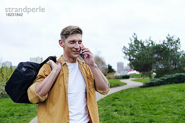 Junger Mann mit Tasche spricht im Park mit Smartphone