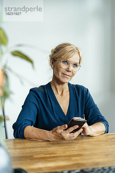 Nachdenkliche Geschäftsfrau mit Brille und Mobiltelefon am Schreibtisch