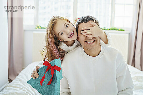 Fröhliches Mädchen hält ein Weihnachtsgeschenk in der Hand und bedeckt die Augen des Vaters  der zu Hause im Bett sitzt