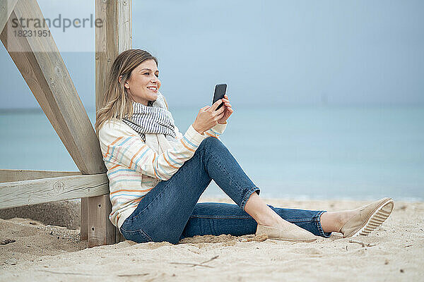 Lächelnde schöne junge Frau  die ihr Smartphone am Strand auf Holz lehnt