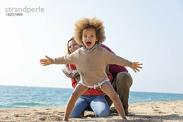 Schreiendes Mädchen steht mit ausgestreckten Armen vor Mutter und Vater am Strand