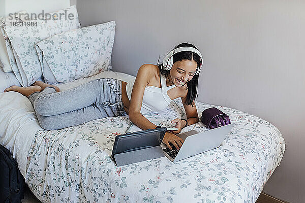 Happy girl with headphones lying on bed using wireless gadgets at home
