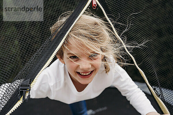 Happy cute girl looking through net on trampoline in garden