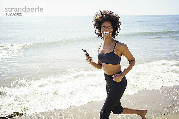 Glückliche junge Frau läuft mit Smartphone am Ufer des Strandes