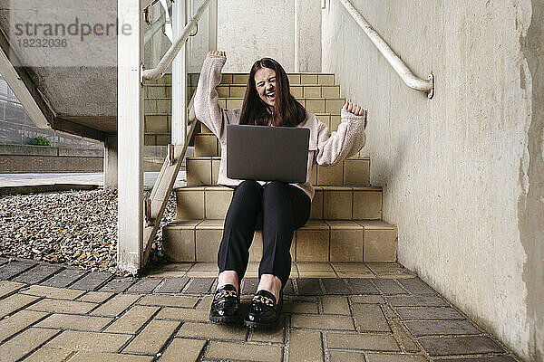 Glückliche junge Frau mit Laptop sitzt auf der Treppe