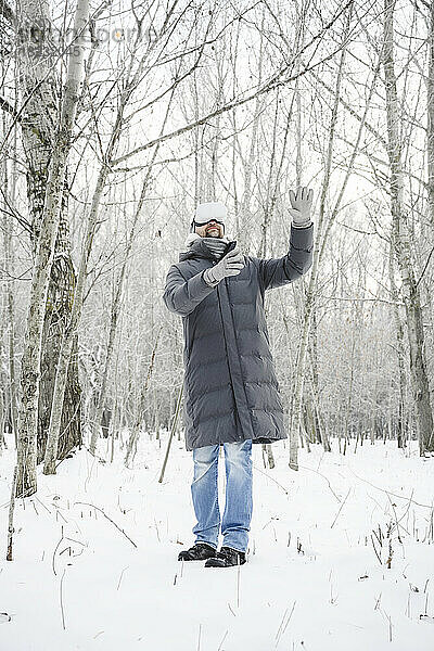 Mann mit Virtual-Reality-Simulator steht auf Schnee und gestikuliert inmitten kahler Bäume